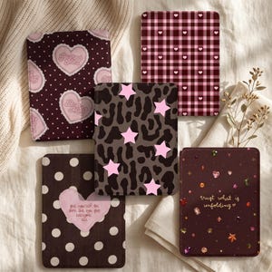 May include: Five rectangular e-reader cases with distinct designs: one with pink hearts on a dark polka dot background, one with a pink plaid heart pattern, one with pink stars on a gray leopard print, one with white polka dots and a heart containing text, and one with gems and the text "trust what is unfolding".