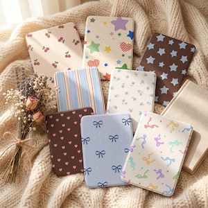 Può includere: Una collezione di quaderni a fantasia con vari disegni, tra cui ciliegie, stelle, cuori, fiocchi e motivi floreali. I quaderni sono disposti su una coperta di maglia color crema, con un bouquet di fiori secchi in primo piano.
