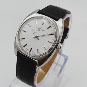 May include: A silver-toned Ulysse Nardin watch with a white face and black leather band. The watch has a round face with silver hands and markers. The band is textured and the watch is displayed on a clear stand.