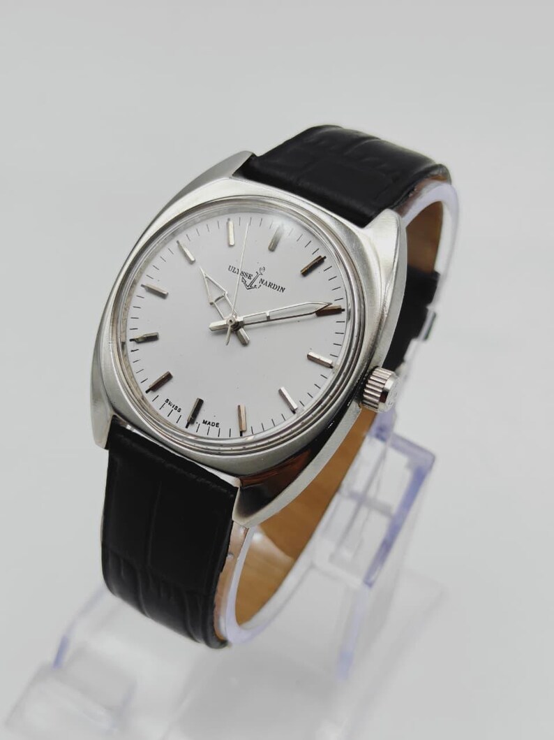 May include: A silver-toned Ulysse Nardin watch with a white face and black leather band. The watch has a round face with silver hands and markers. The band is textured and the watch is displayed on a clear stand.
