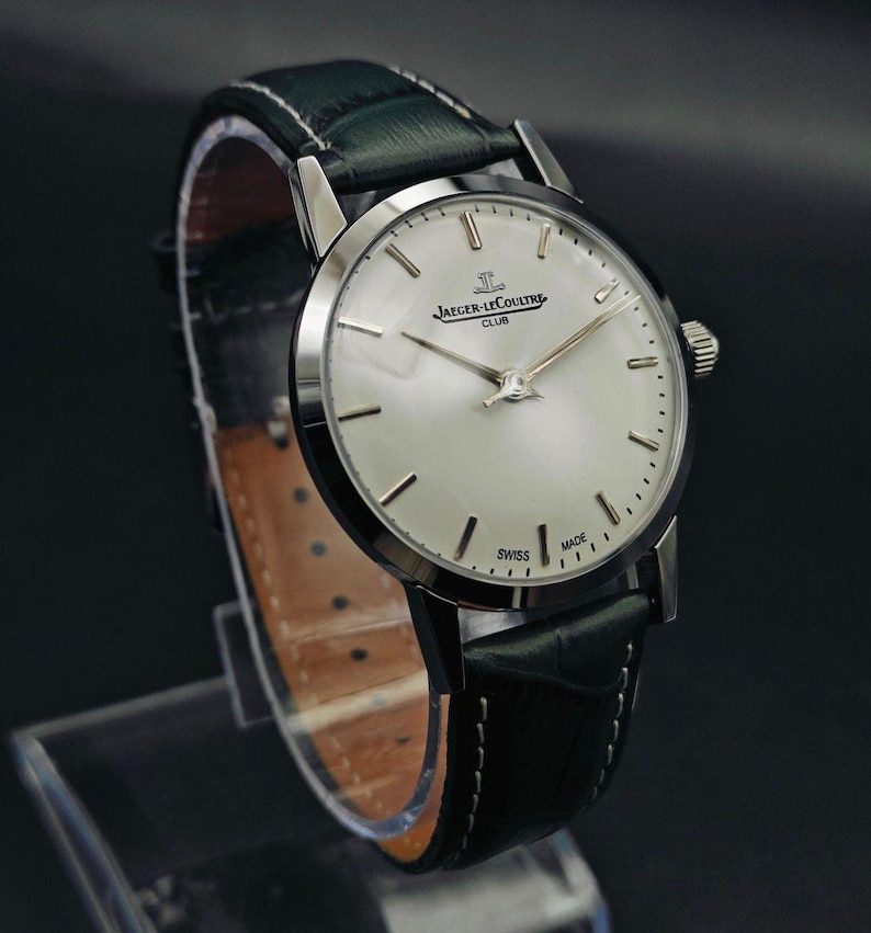 Pu&ograve; includere: Un orologio Jaeger-LeCoultre Club argentato con quadrante chiaro e lancette argentate. L'orologio ha un cinturino in pelle verde scuro ed &egrave; contrassegnato "Swiss Made". L'orologio &egrave; esposto su un supporto trasparente.