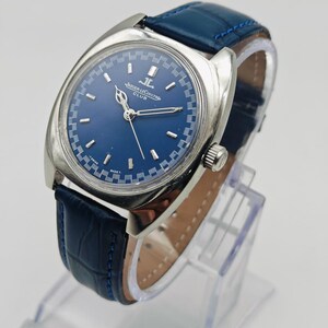 Puede incluir: Un reloj Jaeger-LeCoultre Club vintage con esfera azul y caja plateada. El reloj tiene una correa de cuero azul. El reloj se exhibe en un soporte transparente. La esfera del reloj tiene el logotipo de Jaeger-LeCoultre.