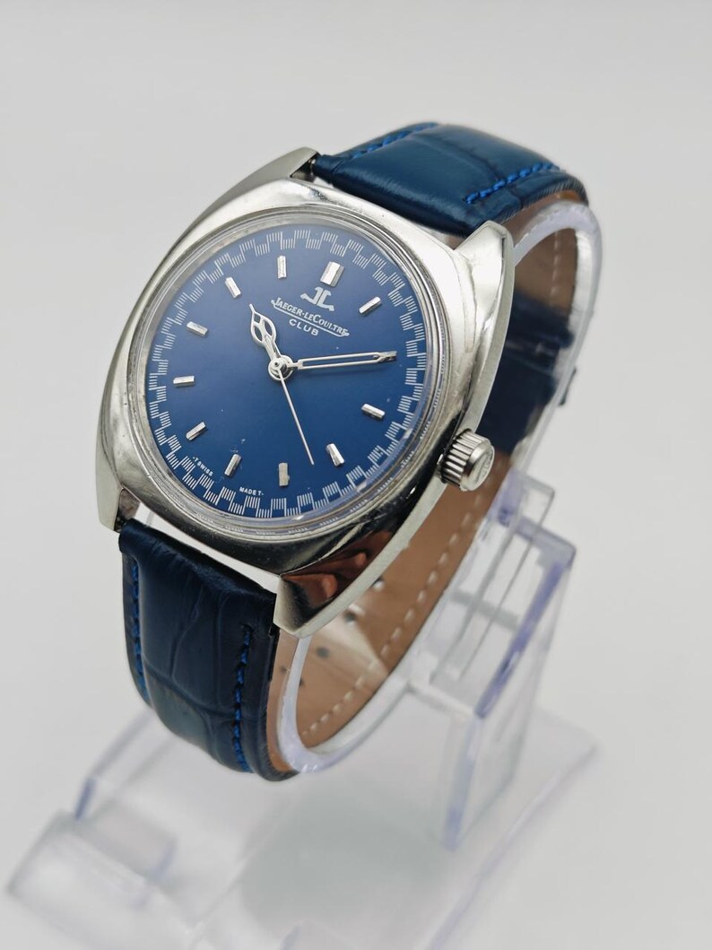 Puede incluir: Un reloj Jaeger-LeCoultre Club vintage con esfera azul y caja plateada. El reloj tiene una correa de cuero azul. El reloj se exhibe en un soporte transparente. La esfera del reloj tiene el logotipo de Jaeger-LeCoultre.