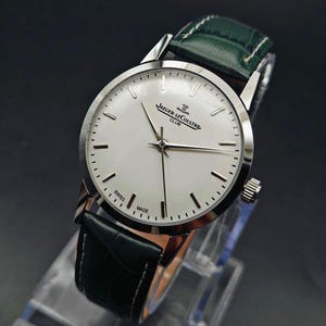 Pu&ograve; includere: Un orologio Jaeger-LeCoultre Club argentato con quadrante bianco e cinturino in pelle nera. L'orologio ha un quadrante rotondo con lancette e indici argentati. Il cinturino &egrave; nero con cuciture verdi. L'orologio &egrave; marchiato "SWISS MADE".