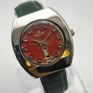 May include: A vintage Breitling Geneve automatic watch with a red dial and a date window. The watch has a silver-tone case and a green leather strap. The watch face features gold-tone hour markers and hands.