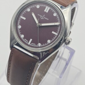 May include: A wristwatch with a burgundy face and a brown leather band. The watch has silver-colored hands and hour markers. The brand name "ULYSSE NARDIN" is displayed on the watch face. The watch is displayed on a clear stand.