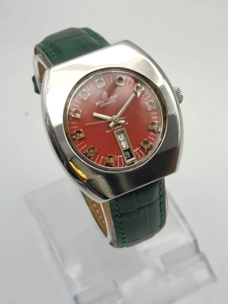 May include: A vintage Breitling Geneve automatic watch with a red dial and a silver-tone case. The watch features a date display and is set on a green leather strap. The watch has a rectangular face.