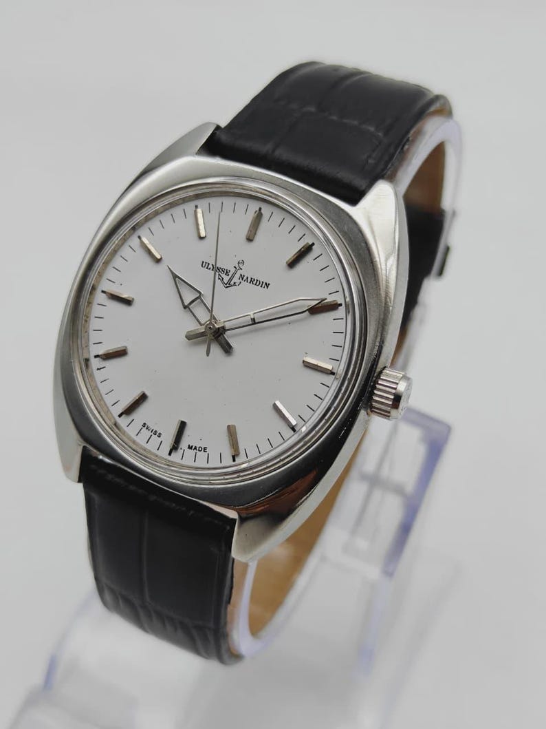 May include: A silver-toned wristwatch with a white face and black leather band. The watch face features black hour markers and the brand name "ULYSSSE NARDIN". The watch is displayed on a clear stand.