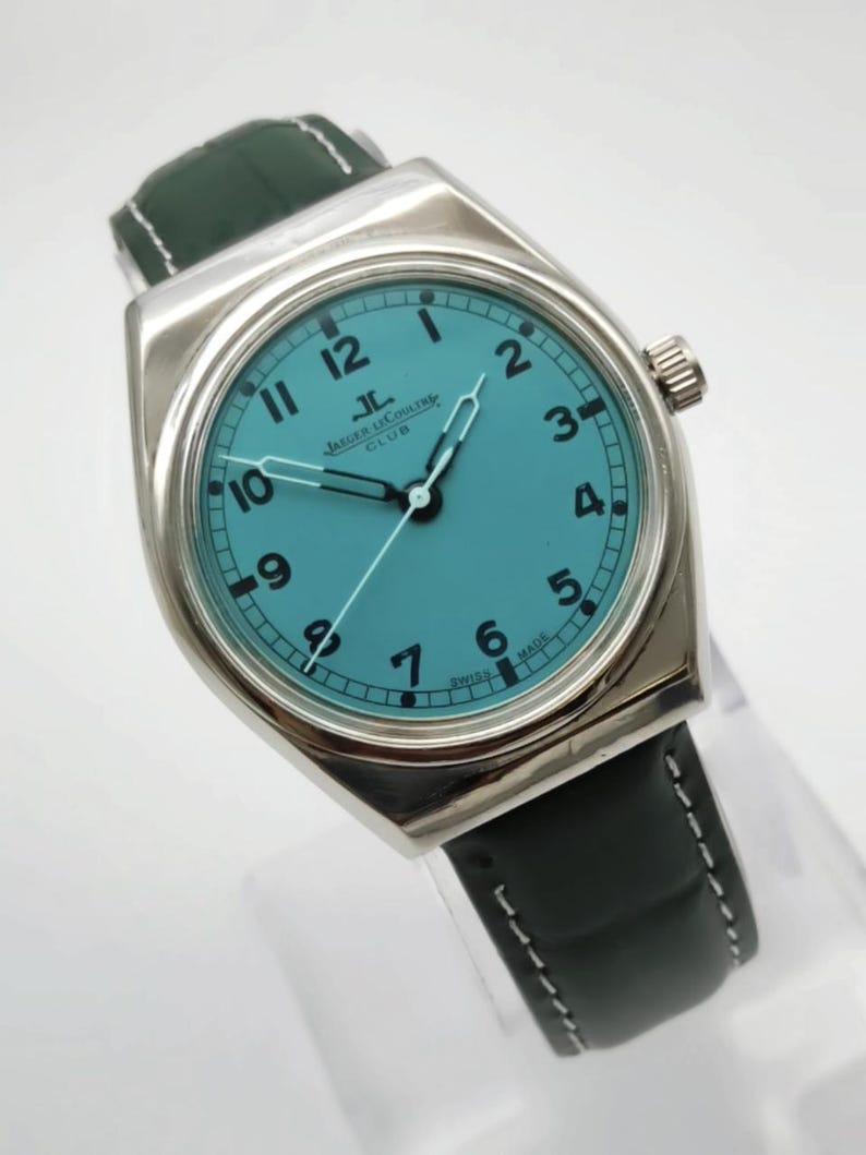 Pode incluir: Um rel&oacute;gio de pulso prateado com mostrador turquesa e algarismos pretos. O rel&oacute;gio apresenta uma bracelete de couro preta com pespontos brancos. O mostrador exibe o log&oacute;tipo Jaeger LeCoultre Club. Um acess&oacute;rio elegante.