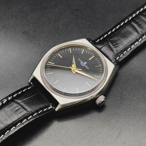 May include: A silver-toned wristwatch with a round black dial, gold-colored hands, and gold-toned hour markers. The watch features the Ulysse Nardin logo and text on the dial. It is attached to a black leather strap with a textured finish and white stitching.