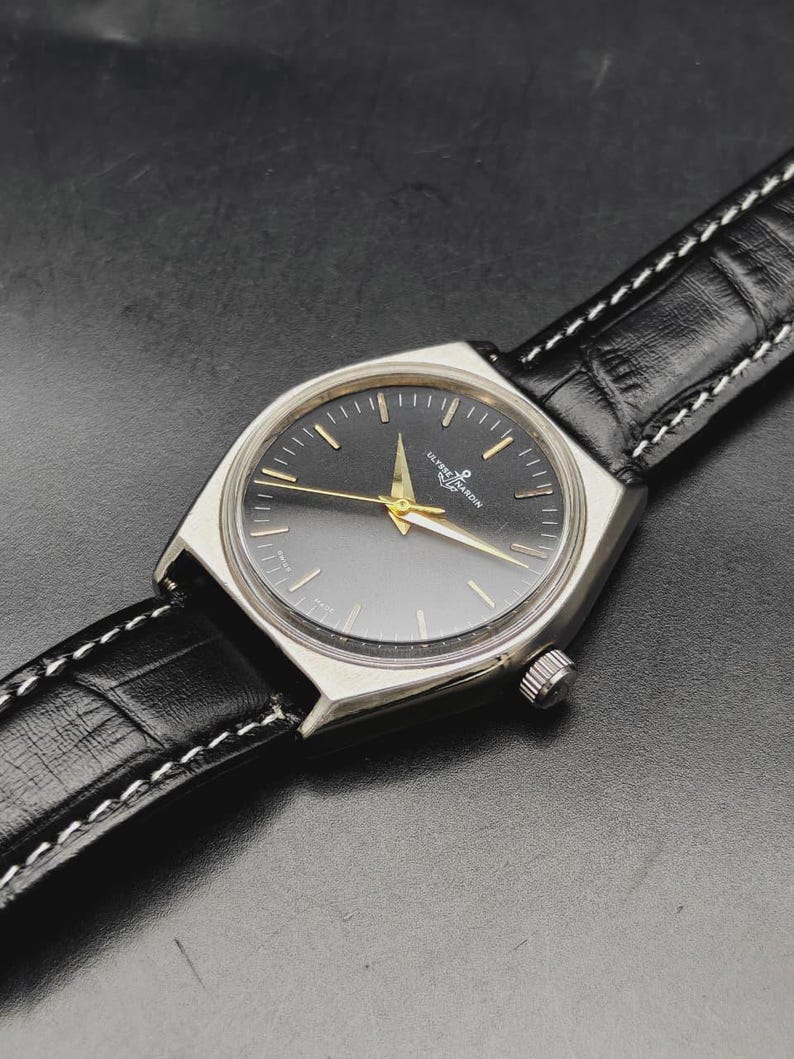 May include: A silver-toned wristwatch with a round black dial, gold-colored hands, and gold-toned hour markers. The watch features the Ulysse Nardin logo and text on the dial. It is attached to a black leather strap with a textured finish and white stitching.