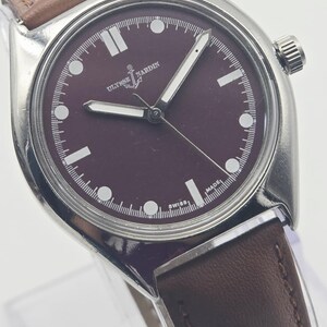 May include: A stainless steel wristwatch with a burgundy dial and a brown leather strap. The watch face features white hour markers and hands, with the brand name "Ulysse Nardin" visible. The watch has a classic, minimalist design.