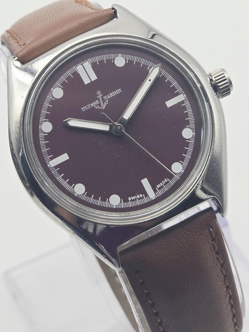 May include: A stainless steel wristwatch with a burgundy dial and a brown leather strap. The watch face features white hour markers and hands, with the brand name "Ulysse Nardin" visible. The watch has a classic, minimalist design.