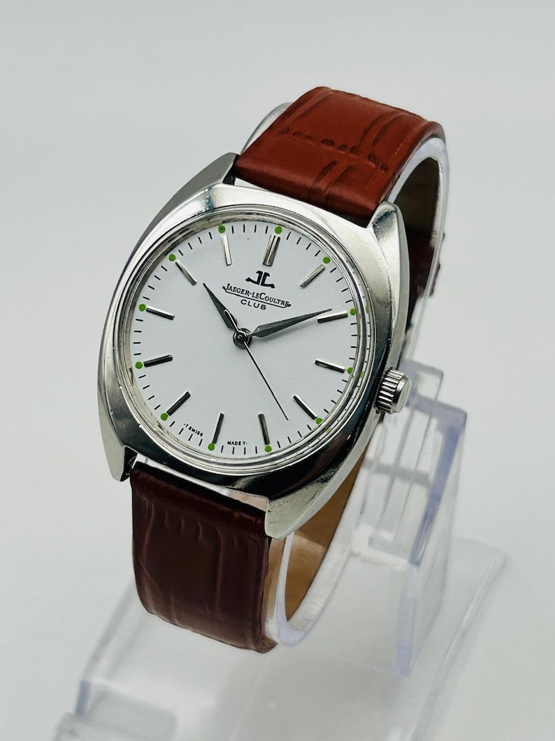 May include: A silver-toned Jaeger-LeCoultre Club watch with a white face and brown leather band. The watch face has black hands and green markers. The band is a rich brown color.