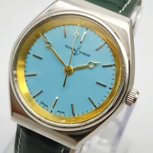 May include: A silver-toned Ulysse Nardin watch with a light blue face and gold-colored hands and markers. The watch has a green leather band and the words "SWISS MADE" are visible on the face.
