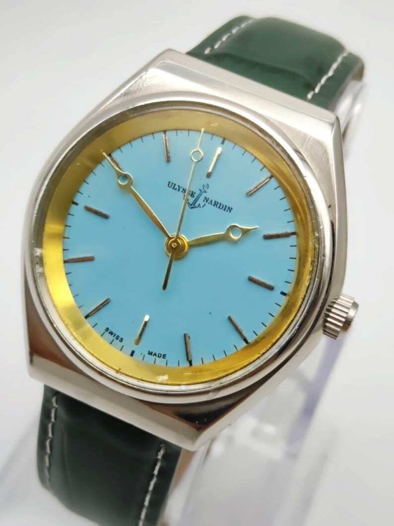 May include: A silver-toned Ulysse Nardin watch with a light blue face and gold-colored hands and markers. The watch has a green leather band and the words "SWISS MADE" are visible on the face.