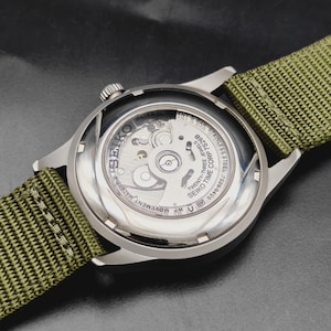 Puede incluir: Un reloj de pulsera de acero inoxidable con una correa de nailon verde oliva. El reloj cuenta con una parte posterior transparente que revela el movimiento mec&aacute;nico interno. El texto incluye SEIKO, TWENTY-THREE JEWELS, 7S26B y 5 BAR.