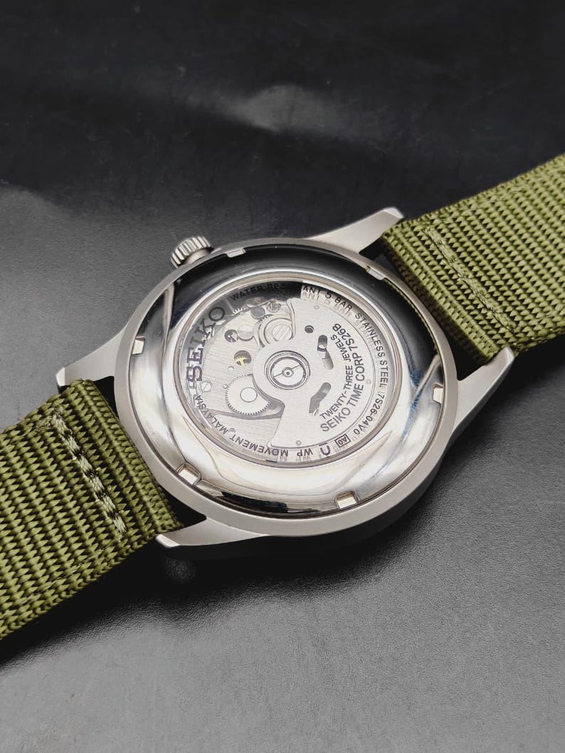 Puede incluir: Un reloj de pulsera de acero inoxidable con una correa de nailon verde oliva. El reloj cuenta con una parte posterior transparente que revela el movimiento mec&aacute;nico interno. El texto incluye SEIKO, TWENTY-THREE JEWELS, 7S26B y 5 BAR.
