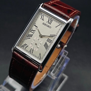 May include: A rectangular Seiko wristwatch with a silver-colored face, black Roman numerals, and a subdial. The watch has a brown leather band. The watch is displayed on a clear stand against a dark background.