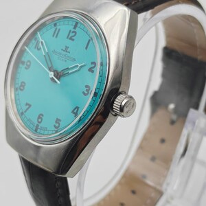 Op de afbeelding: Een zilverkleurig polshorloge met een turquoise wijzerplaat en een zwarte leren band. De wijzerplaat heeft zwarte cijfers en wijzers, met de merknaam "Jaeger-LeCoultre Club". Het horloge wordt op een transparante standaard getoond.