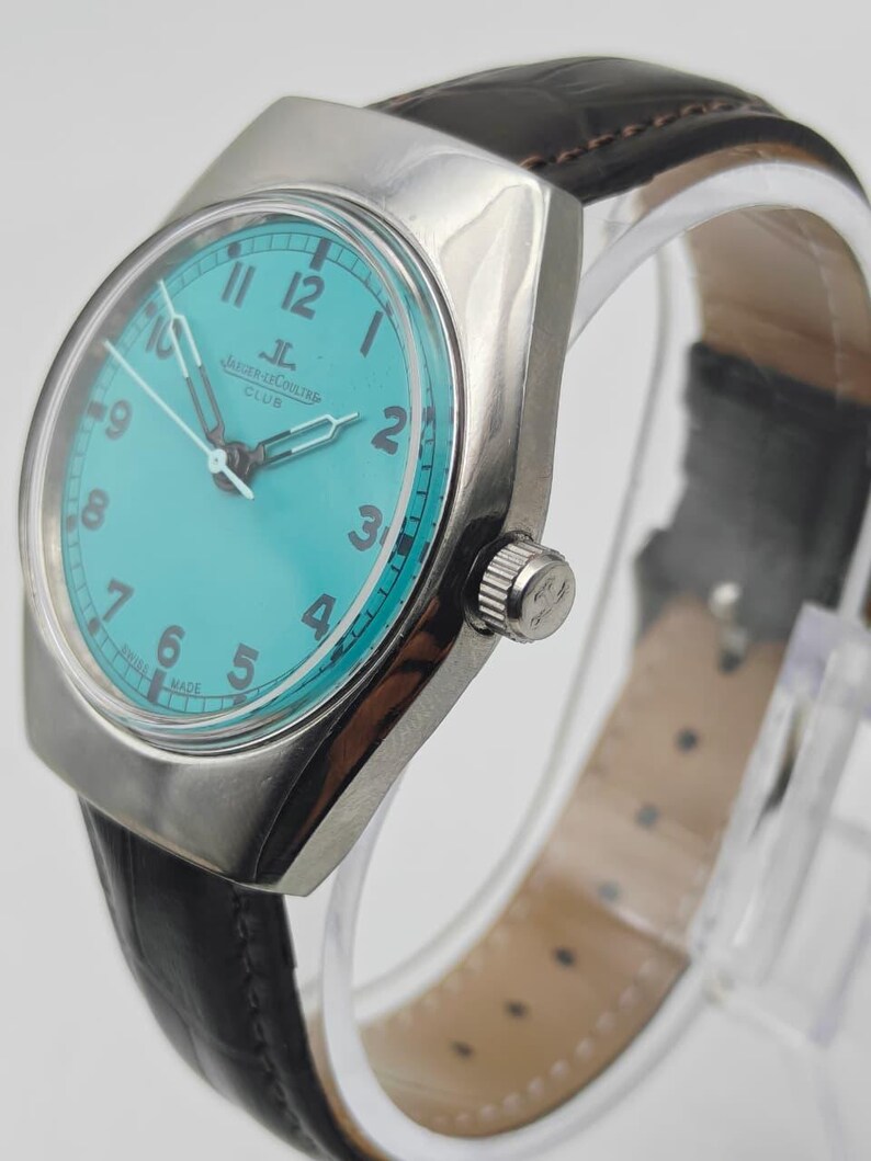 Op de afbeelding: Een zilverkleurig polshorloge met een turquoise wijzerplaat en een zwarte leren band. De wijzerplaat heeft zwarte cijfers en wijzers, met de merknaam "Jaeger-LeCoultre Club". Het horloge wordt op een transparante standaard getoond.