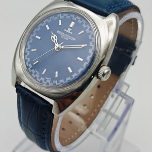 Puede incluir: Un reloj Jaeger-LeCoultre Club plateado con esfera azul y correa de cuero azul. La esfera del reloj presenta marcadores de hora y agujas plateadas. La correa est&aacute; cosida con hilo azul.