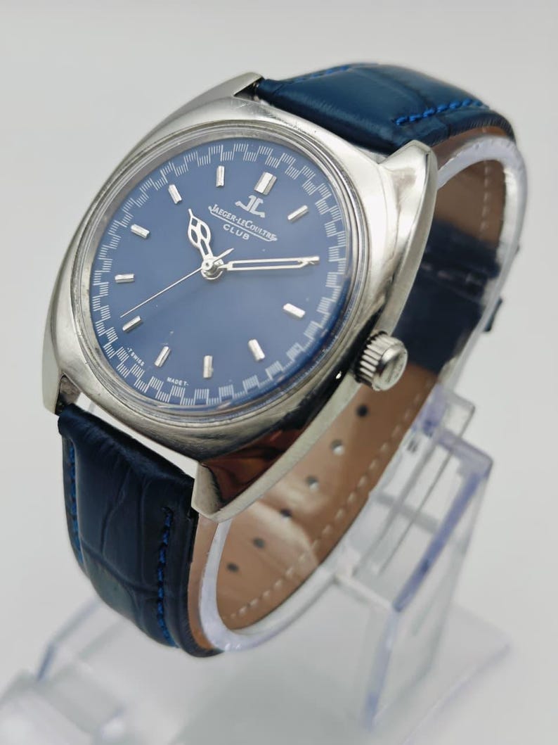 Puede incluir: Un reloj Jaeger-LeCoultre Club plateado con esfera azul y correa de cuero azul. La esfera del reloj presenta marcadores de hora y agujas plateadas. La correa est&aacute; cosida con hilo azul.