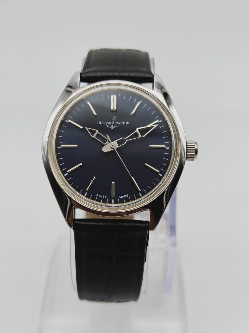 May include: A silver-toned Ulysse Nardin wristwatch with a dark blue face and black leather band. The watch features a round face with silver hands and markers. The text "SWISS MADE" is visible on the watch face.