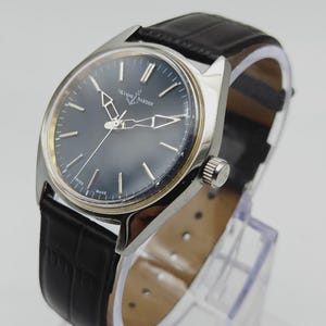 May include: A silver-toned Ulysse Nardin wristwatch with a dark blue face and black leather band. The watch features silver hands and markers, and the band has a crocodile pattern. The watch is displayed on a clear stand.