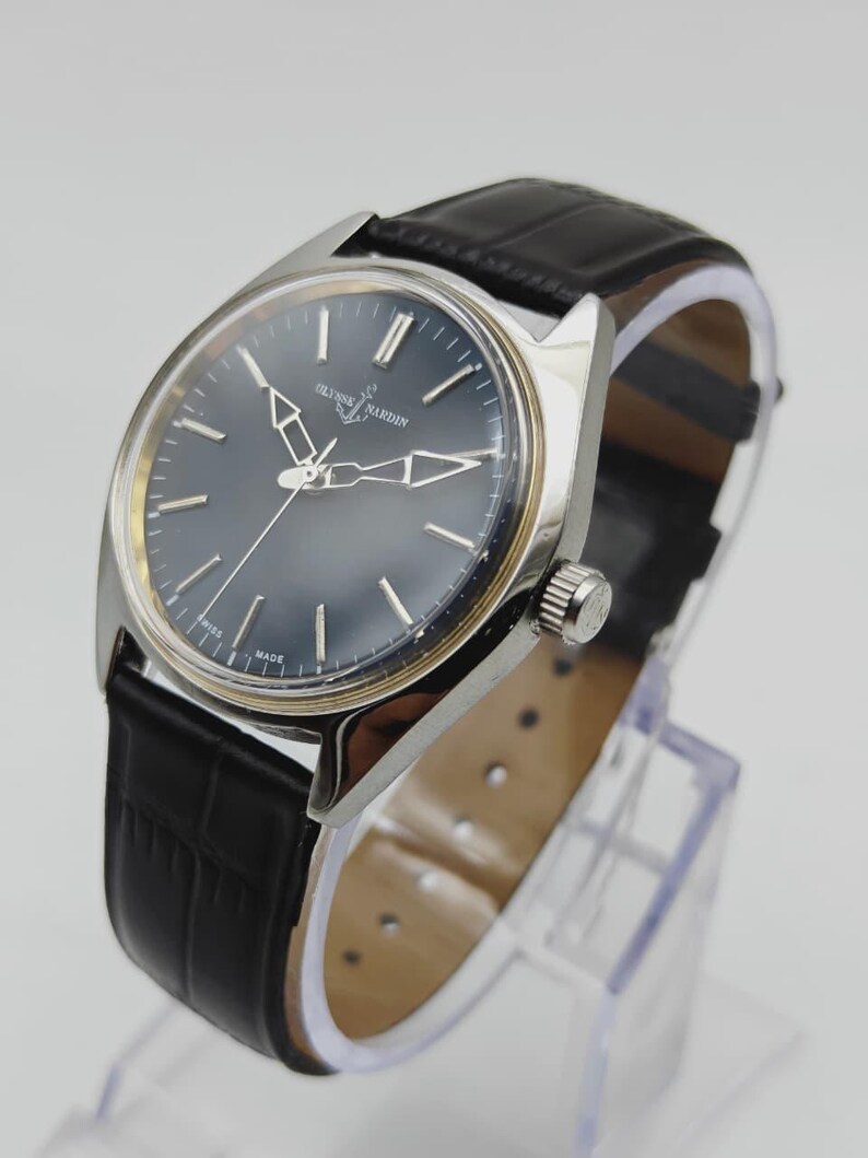 May include: A silver-toned Ulysse Nardin wristwatch with a dark blue face and black leather band. The watch features silver hands and markers, and the band has a crocodile pattern. The watch is displayed on a clear stand.