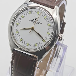May include: A silver-toned Jaeger-LeCoultre Club watch with a white face, featuring the brand's logo and the words "SWISS MADE." The watch has a brown leather band with white stitching and gold-toned hands.
