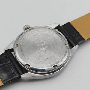 May include: Close-up of a vintage wristwatch, showcasing the textured silver-toned back casing and crown. The watch features a black leather band with a light brown underside. The watch's design suggests a classic, timeless aesthetic.