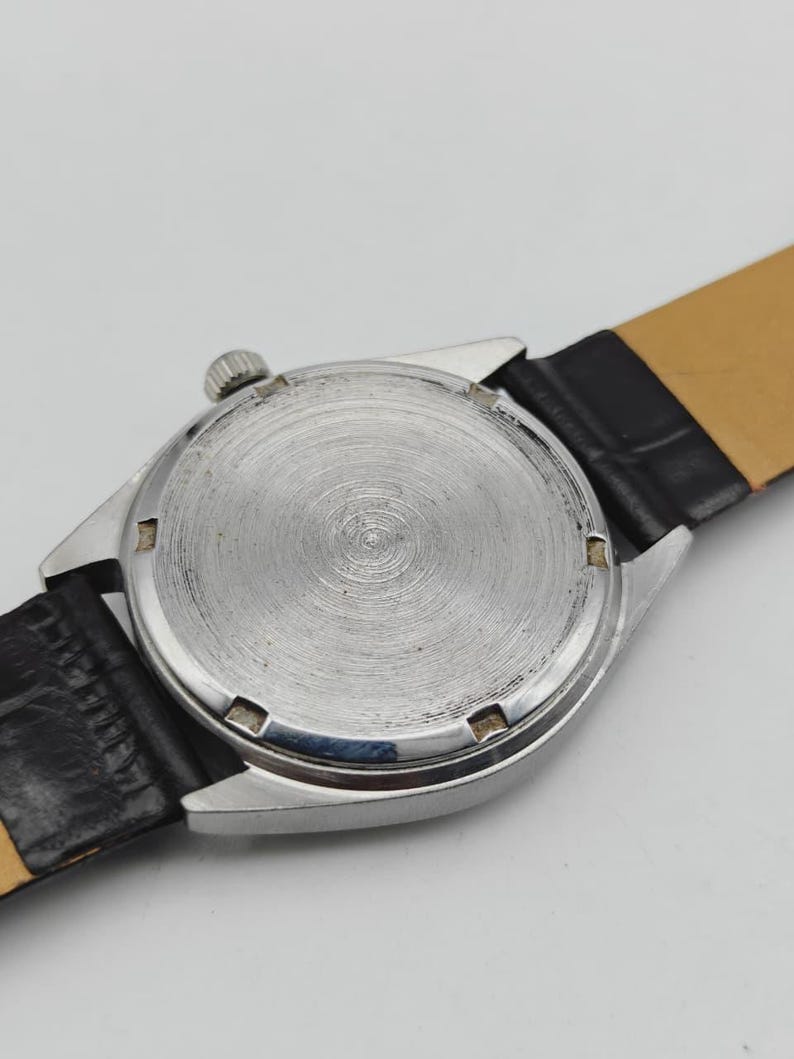 May include: Close-up of a vintage wristwatch, showcasing the textured silver-toned back casing and crown. The watch features a black leather band with a light brown underside. The watch's design suggests a classic, timeless aesthetic.