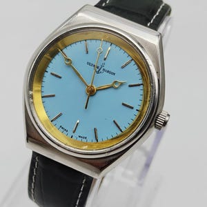 May include: A vintage Ulysse Nardin watch with a light blue face and gold-tone hands and markers. The watch has a silver-tone case and a black leather band with white stitching. The watch face has the text "Ulysse Nardin" and "Swiss Made".