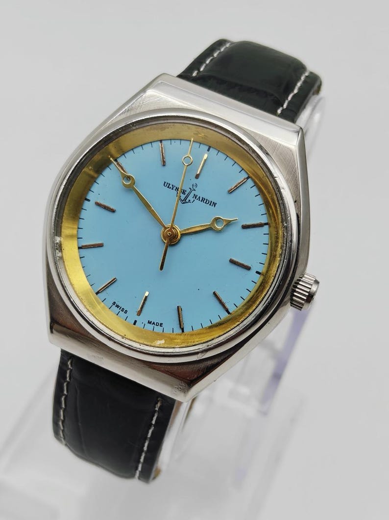 May include: A vintage Ulysse Nardin watch with a light blue face and gold-tone hands and markers. The watch has a silver-tone case and a black leather band with white stitching. The watch face has the text "Ulysse Nardin" and "Swiss Made".