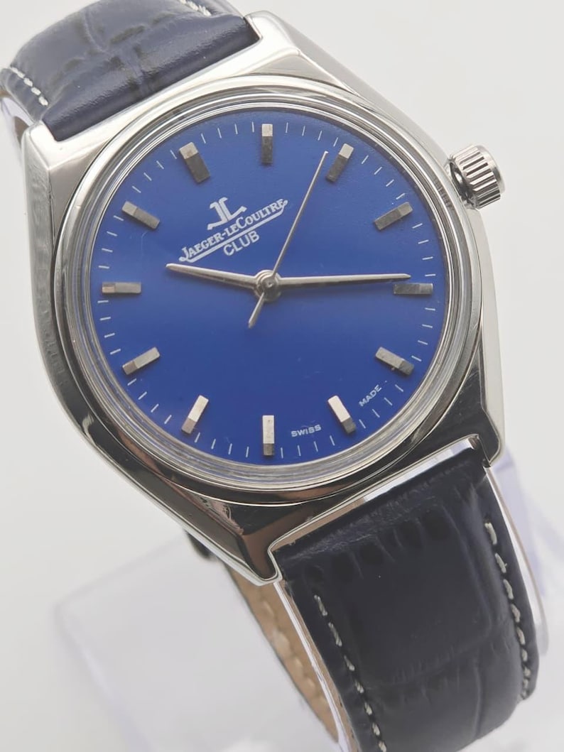May include: A silver-toned wristwatch with a deep blue face and a black leather band. The watch face features silver-toned markers and hands, with the Jaeger-LeCoultre Club logo. The watch has a textured crown and is marked "Swiss Made".