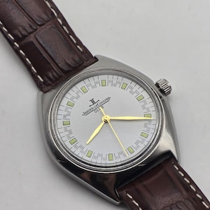 May include: A silver-toned Jaeger-LeCoultre Club watch with a light-colored face, gold hands, and a brown leather strap. The watch has a rectangular case and a textured brown leather band with white stitching.