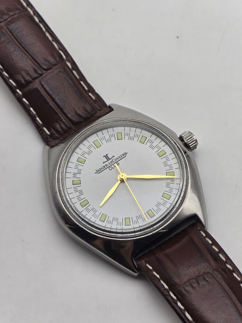 May include: A silver-toned Jaeger-LeCoultre Club watch with a light-colored face, gold hands, and a brown leather strap. The watch has a rectangular case and a textured brown leather band with white stitching.