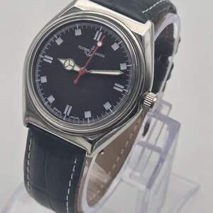 Puede incluir: Un reloj Ulysse Nardin de acero inoxidable con esfera negra y correa de cuero negro. El reloj tiene un bisel plateado, marcadores blancos y un segundero rojo. La correa tiene costuras blancas.