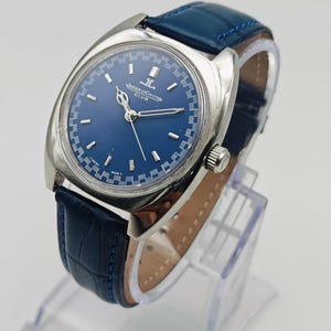 Puede incluir: Un reloj Jaeger-LeCoultre Club vintage con esfera azul y caja plateada. El reloj tiene una correa de cuero azul. El reloj se exhibe en un soporte transparente. La esfera del reloj tiene el logotipo de Jaeger-LeCoultre.