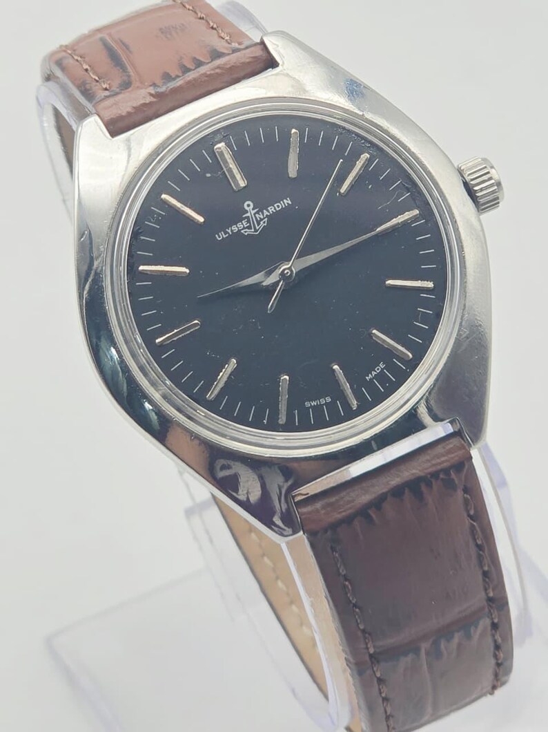 May include: A silver-toned Ulysse Nardin wristwatch with a black face and brown leather band. The watch face features silver hour markers and hands. The brand name and anchor logo are visible on the watch face. The watch is marked "Swiss Made".