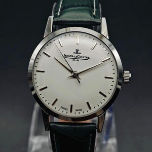 Pu&ograve; includere: Un orologio Jaeger-LeCoultre Club argentato con quadrante bianco, lancette nere e cinturino in pelle nera. Il quadrante mostra il logo Jaeger-LeCoultre e le parole "SWISS MADE". L'orologio ha un cinturino in pelle verde.