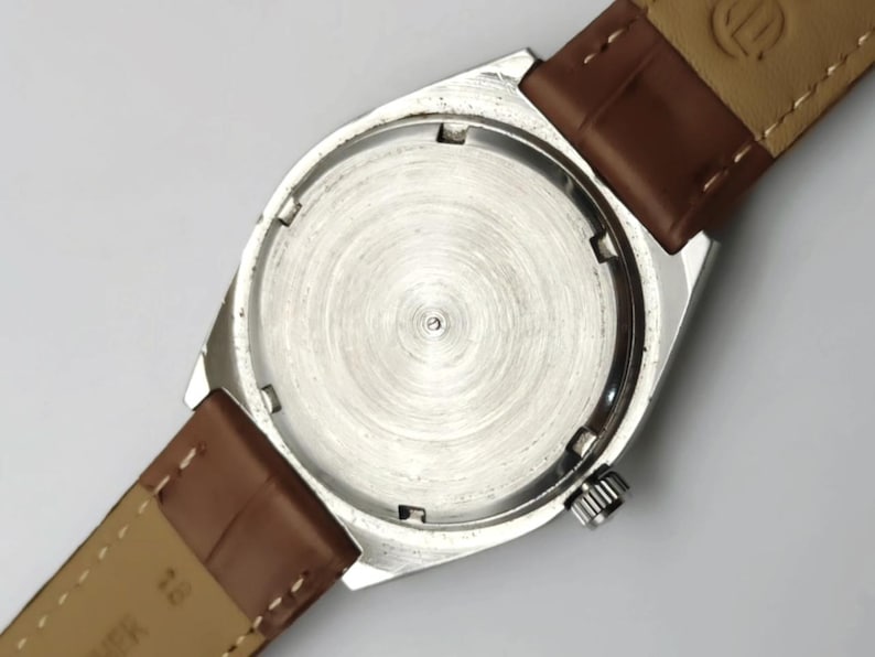 K&ouml;nnte beinhalten: Eine silberfarbene Armbanduhr mit braunem Lederarmband. Das Uhrengeh&auml;use ist rechteckig mit rundem Boden. Das Armband ist mit Federstegen am Geh&auml;use befestigt. Die Uhr hat eine silberfarbene Krone.