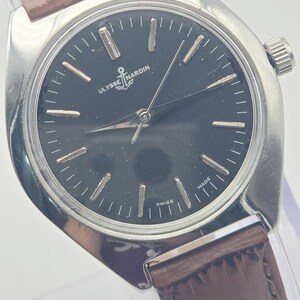 May include: A silver-toned Ulysse Nardin wristwatch with a dark face and brown leather band. The watch face features silver hour markers and hands, with the Ulysse Nardin logo and the word "SWISS MADE". The watch has a classic, elegant design.