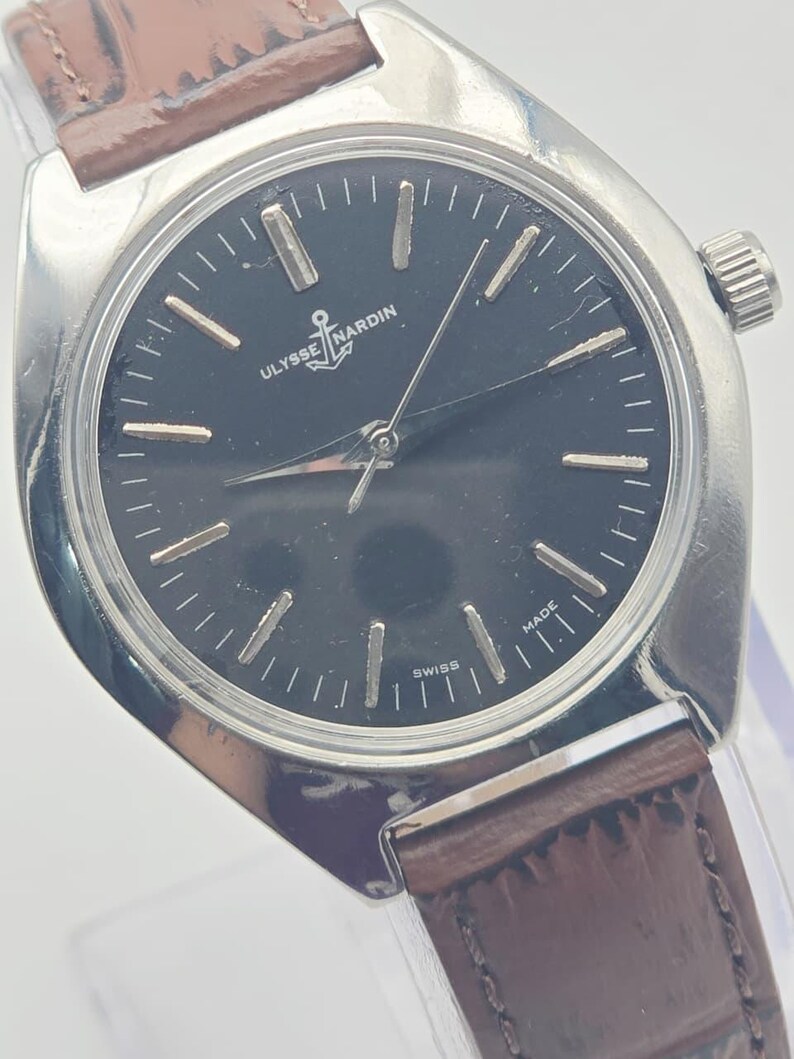 May include: A silver-toned Ulysse Nardin wristwatch with a dark face and brown leather band. The watch face features silver hour markers and hands, with the Ulysse Nardin logo and the word "SWISS MADE". The watch has a classic, elegant design.