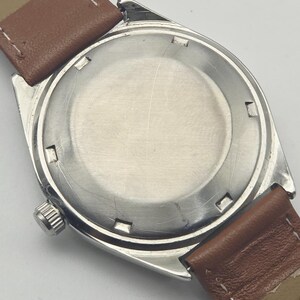 May include: Close-up of the back of a vintage wristwatch. The watch has a silver-colored case with a round, brushed metal back. Brown leather watch straps are attached. The watch's crown is visible on the side.