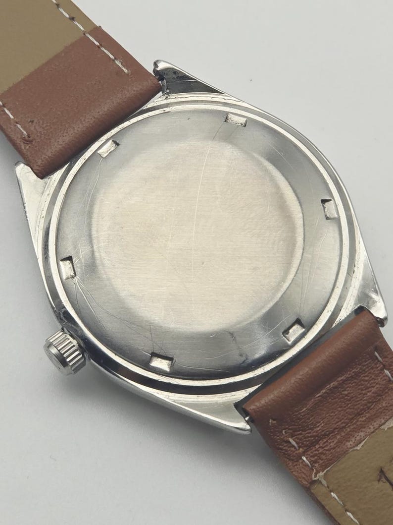 May include: Close-up of the back of a vintage wristwatch. The watch has a silver-colored case with a round, brushed metal back. Brown leather watch straps are attached. The watch's crown is visible on the side.