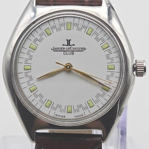 May include: A silver-toned Jaeger-LeCoultre Club watch with a white face, gold hands, and a brown leather strap. The watch face features the brand name and the words "SWISS MADE". The watch has a round face with rectangular markers.