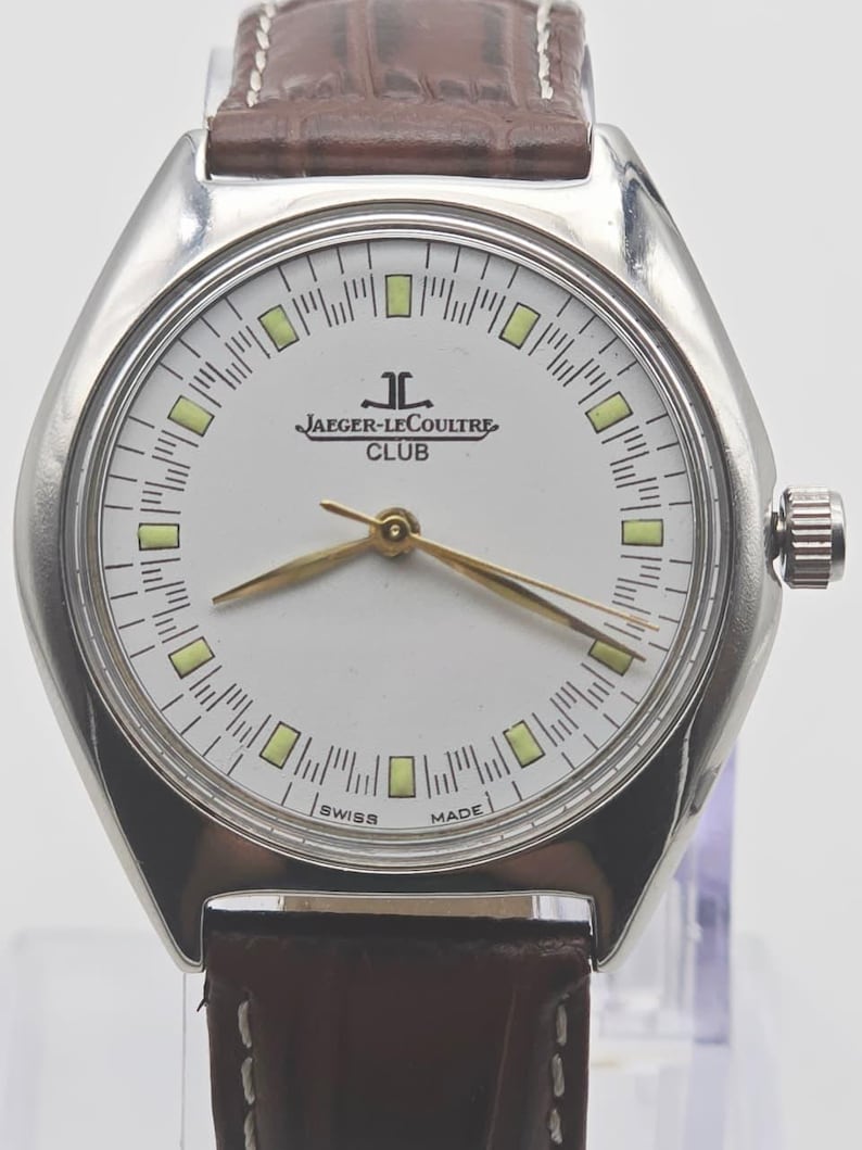May include: A silver-toned Jaeger-LeCoultre Club watch with a white face, gold hands, and a brown leather strap. The watch face features the brand name and the words "SWISS MADE". The watch has a round face with rectangular markers.