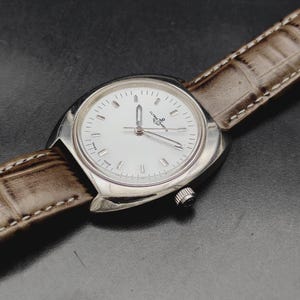 May include: A silver-toned wristwatch with a white dial, silver hour markers, and silver hands. The watch features the Ulysse Nardin logo on the face and is attached to a textured, light brown leather strap with visible white stitching along the edges.